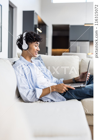 Wearing headphones, teenage boy relaxing on couch and using laptop at home 116315072