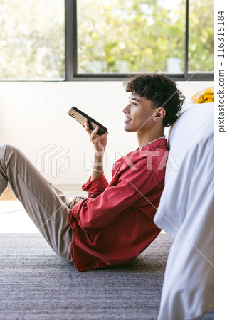 Sitting on floor, young man using smartphone and smiling in cozy home setting Sitting on floor, young man using smartphone and smiling in cozy home setting 116315184