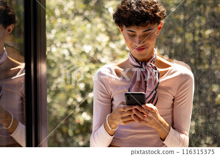 Texting on smartphone, transgender woman standing by window, smiling and relaxed Texting on smartphone, transgender woman standing by window, smiling and relaxed 116315375