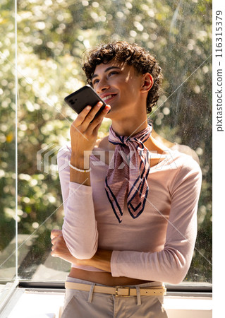 Smiling and holding smartphone, transgender woman enjoying conversation by window 116315379