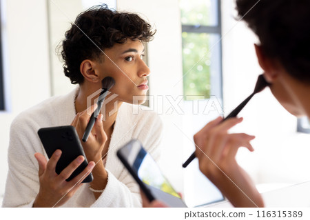 Applying makeup, transgender woman using smartphone while looking in mirror 116315389
