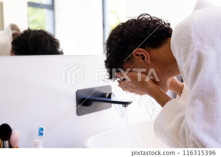 Washing face in bathroom, young man wearing white robe in front of mirror 116315396