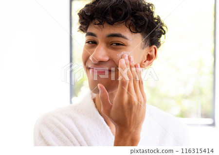 Applying face cream, young young man in bathrobe smiling in natural light Applying face cream, young young man in bathrobe smiling in natural light 116315414