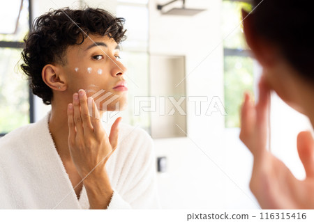 Applying skincare cream, teenage boy in white robe looking in mirror 116315416