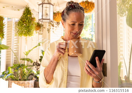 Holding smartphone and drinking coffee, mature woman relaxing at home Holding smartphone and drinking coffee, mature woman relaxing at home 116315516