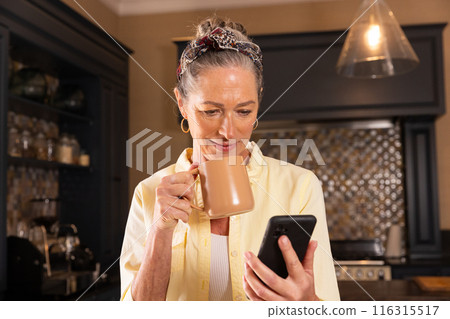 Holding mug, mature woman reading smartphone message in kitchen Holding mug, mature woman reading smartphone message in kitchen 116315517