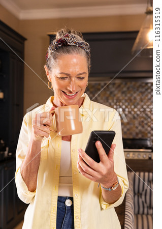 Holding coffee mug, senior woman smiling while reading on smartphone in kitchen 116315519