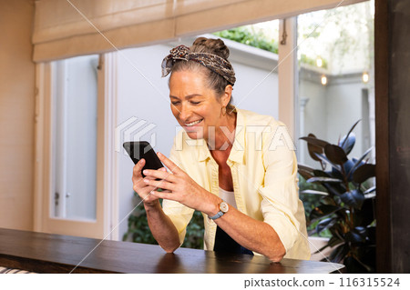 Smiling senior woman using smartphone while leaning on table at home 116315524