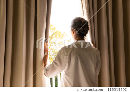 Opening curtains, mature woman in white shirt looking outside window at home, copy space Opening curtains, mature woman in white shirt looking outside window at home, copy space 116315592