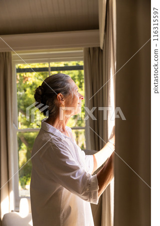Opening curtains, mature woman enjoying sunlight at home, looking outside 116315597