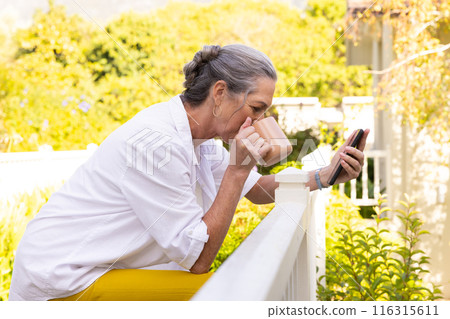 Drinking coffee, mature woman using smartphone while leaning on balcony railing 116315611