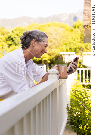 Smiling mature woman using smartphone and drinking coffee on porch railing Smiling mature woman using smartphone and drinking coffee on porch railing 116315613