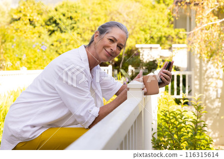 Smiling mature woman holding smartphone and coffee cup, relaxing on porch 116315614