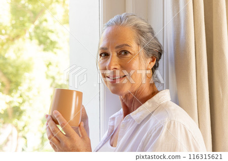 Smiling mature woman holding coffee cup standing by window at home Smiling mature woman holding coffee cup standing by window at home 116315621