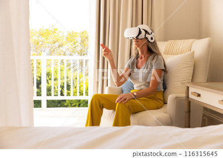 Using VR headset, mature woman sitting on chair interacting with virtual environment 116315645