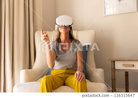 Using VR headset, mature woman sitting on chair interacting with virtual environment Using VR headset, mature woman sitting on chair interacting with virtual environment 116315647