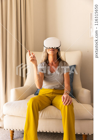 Using VR headset, mature woman sitting on chair interacting with virtual environment 116315650