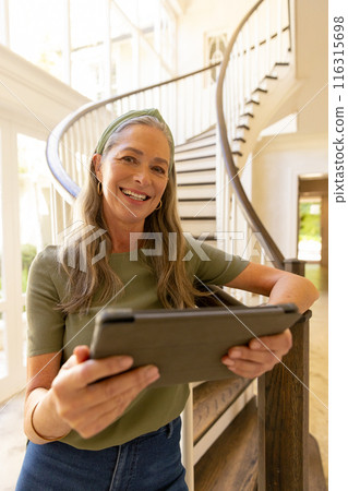 Smiling woman using tablet while standing near staircase at home 116315698