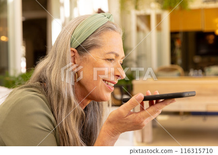Smiling mature woman using smartphone for voice message at home, enjoying technology 116315710