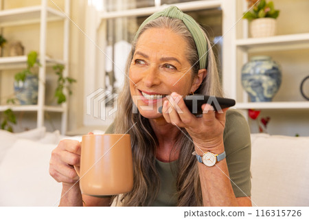 Holding smartphone and drinking coffee, mature woman smiling and relaxing at home Holding smartphone and drinking coffee, mature woman smiling and relaxing at home 116315726