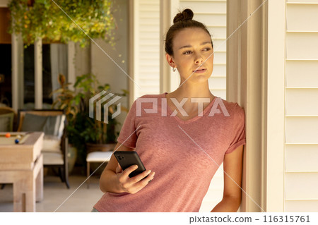 Holding smartphone, young woman leaning against wall and looking away thoughtfully 116315761