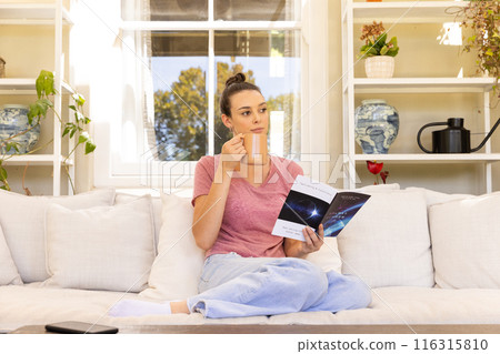 Sitting on couch, woman drinking coffee and reading book in living room Sitting on couch, woman drinking coffee and reading book in living room 116315810