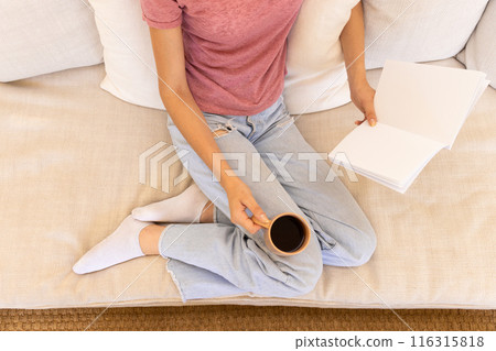 Reading book and holding coffee cup, woman relaxing on couch at home 116315818
