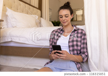 Using smartphone, young woman sitting on floor in bedroom, relaxing 116315851