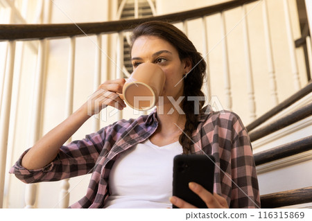 Drinking coffee and using smartphone, woman relaxing on staircase at home Drinking coffee and using smartphone, woman relaxing on staircase at home 116315869