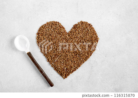 A buckwheat in the shape of a heart and ceramic spoon as a symbol of good for the heart. Buckwheat groats on a light background. copy space 116315870
