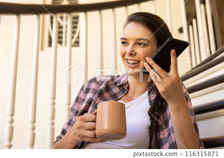 Holding smartphone and coffee mug, woman smiling and talking at home 116315871