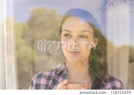 Young woman holding coffee cup, looking outside window, enjoying peaceful moment 116315874