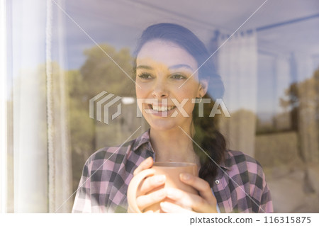 Smiling woman holding coffee cup, looking out window at home, feeling relaxed 116315875