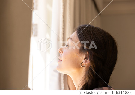 Smiling woman looking out window, enjoying sunlight and peaceful moment at home, copy space Smiling woman looking out window, enjoying sunlight and peaceful moment at home, copy space 116315882
