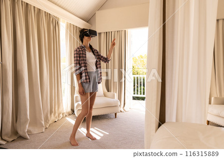 Using VR headset, young woman exploring virtual reality in living room Using VR headset, young woman exploring virtual reality in living room 116315889