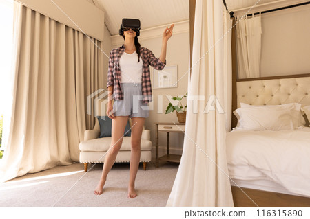 Using VR headset, woman standing in bedroom and pointing at something Using VR headset, woman standing in bedroom and pointing at something 116315890