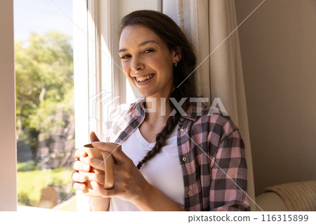 Smiling young woman holding coffee cup and standing by window at home 116315899