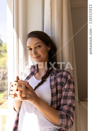 Smiling woman holding coffee cup, standing by window at home Smiling woman holding coffee cup, standing by window at home 116315915