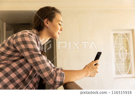Using smartphone, young woman leaning on railing and smiling at home 116315916