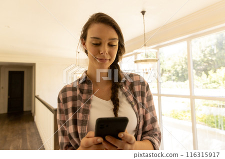 Using smartphone, young woman in casual clothes standing in bright home 116315917
