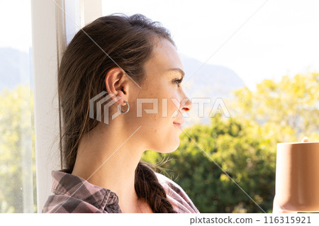 Smiling woman holding coffee cup, looking out window at nature 116315921