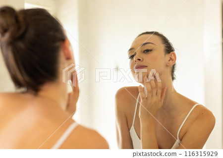 Checking skin in mirror, young woman focusing on skincare routine at home Checking skin in mirror, young woman focusing on skincare routine at home 116315929