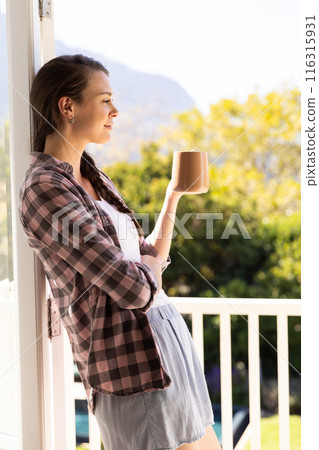 Young woman enjoying morning coffee, standing on balcony and looking outside 116315931