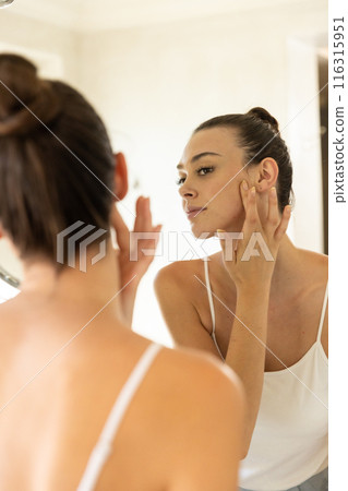 Applying skincare, young woman examining her face in bathroom mirror Applying skincare, young woman examining her face in bathroom mirror 116315951