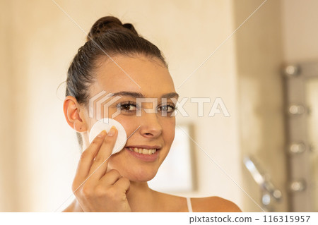Smiling woman applying facial cleanser with cotton pad in bathroom 116315957