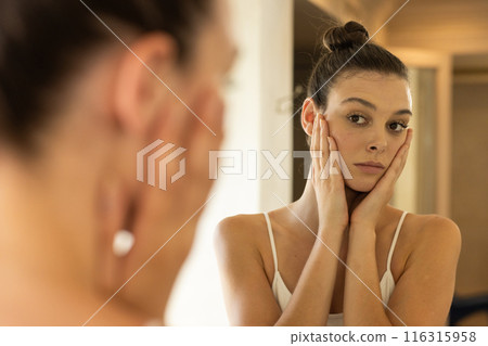 Looking in mirror, young woman touching face with thoughtful expression 116315958