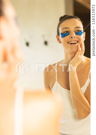 Applying under-eye patches, woman smiling and looking in mirror at home Applying under-eye patches, woman smiling and looking in mirror at home 116315972