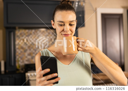 Holding smartphone and drinking coffee, woman enjoying morning in kitchen 116316026