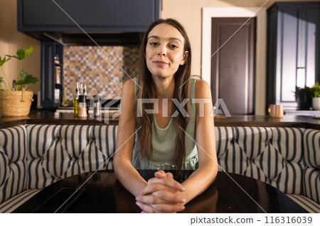 Smiling young woman sitting at kitchen table with hands clasped Smiling young woman sitting at kitchen table with hands clasped 116316039