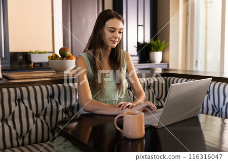 Working from home, young woman using laptop and drinking coffee in kitchen Working from home, young woman using laptop and drinking coffee in kitchen 116316047